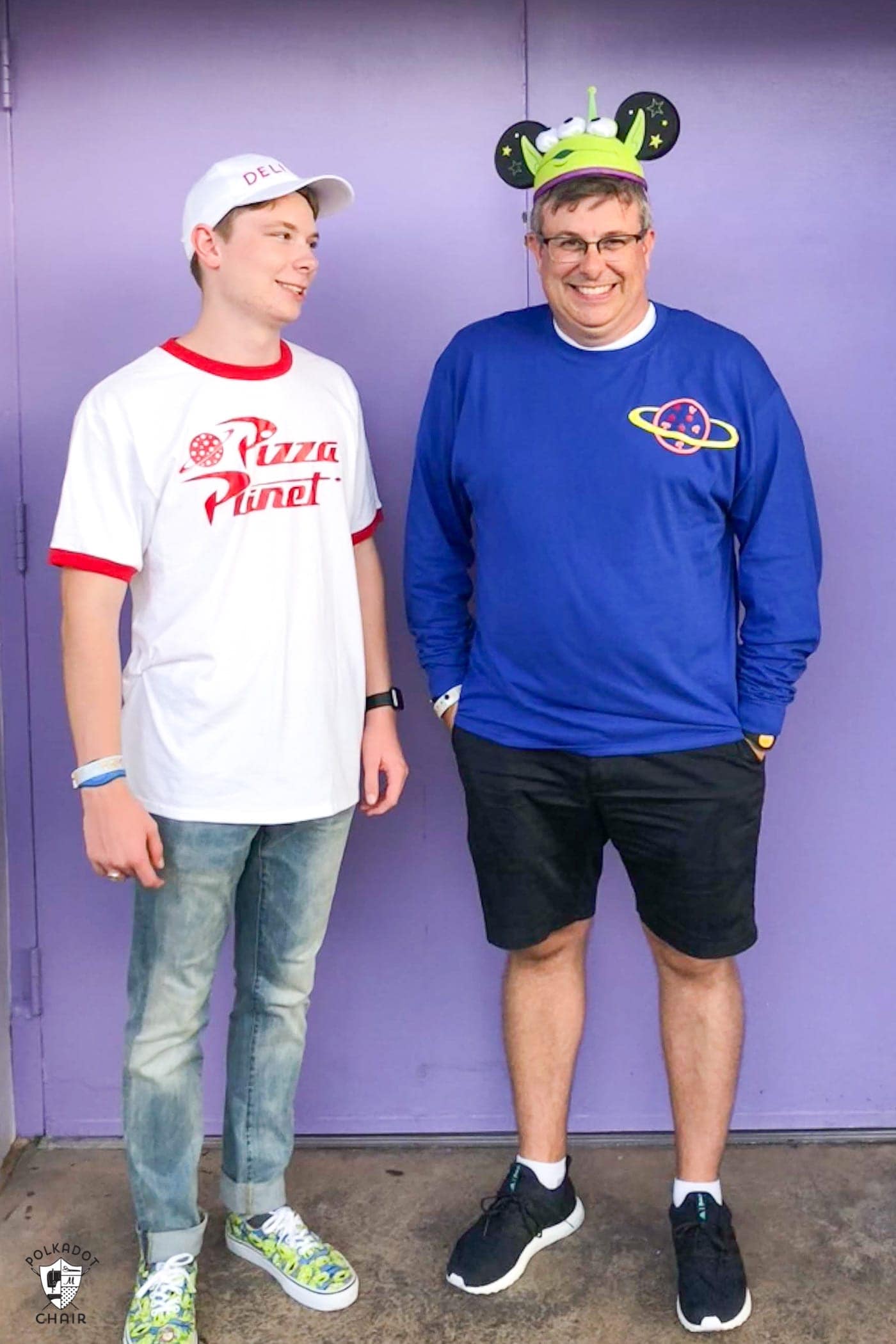 two men standing in front of a purple wall. One is in a white baseball cap and wearing a white tshirt with red lining the collar and sleeve with the Pizza Planet logo. The other is in a dark blue long sleeve tshirt with a pizza surrounded by a ring in the top corner. and has on a green alien mickey hat. 